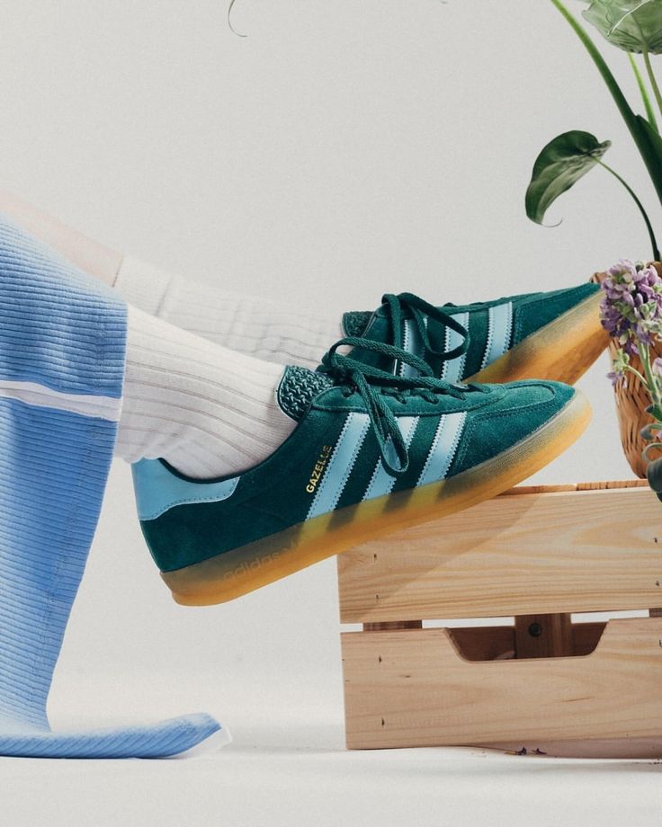 Chaussures Adidas Gazelle Indoor Collegiate Green portées. Design vert foncé et bleu clair avec semelles gomme. Chaussettes blanches. Fond avec des plantes et une caisse en bois. Style décontracté et moderne.