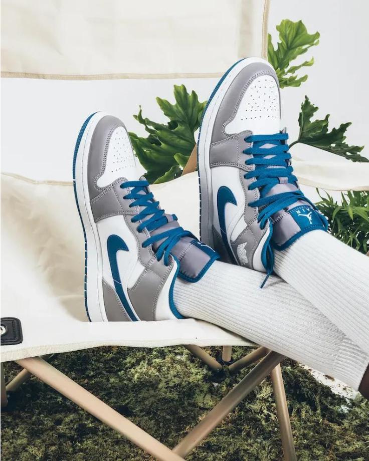 Chaussures Air Jordan 1 Mid True Blue portées sur une chaise beige. Design blanc, gris et bleu avec lacets bleus. Chaussettes blanches. Fond naturel avec plantes vertes. Style décontracté et tendance.